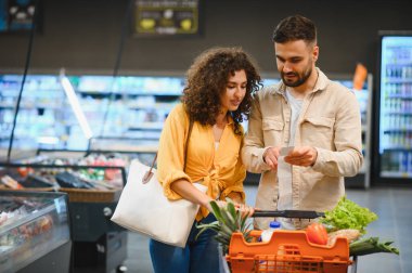 Alışveriş listesine bakan genç bir çift, alışveriş mağazasında içi taze yiyecek ve bakkaliyeyle dolu bir arabayı itiyorlar.