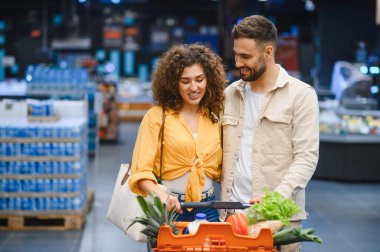 Gülümseyen, içi taze yiyeceklerle dolu bir alışveriş arabasını iten genç bir çift, aydınlık bir market koridorunda yürürken...