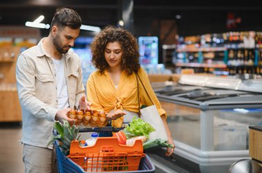 Çift birlikte alışveriş yapıyor, süpermarkette yumurtalara bakıyor. Yumurta kutusu tutan bir adam, alışveriş arabasında eşyalar gören bir kadın.