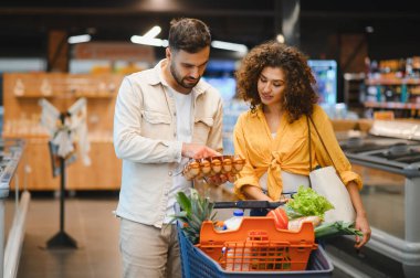 Genç bir çift markette ürünleri karşılaştırıyor, taze ürünlerle dolu alışveriş arabalarını itiyor, sağlıklı gıda seçimleri yapıyor.