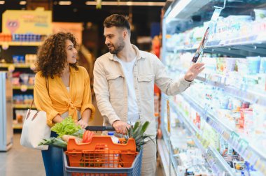 Buzdolabından sağlıklı süt ürünleri seçen genç bir çift, içi taze ürünlerle dolu bir alışveriş arabasını itiyor.