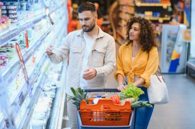 Genç çift mandıra ürünlerine bakarken markette içi taze ürünlerle dolu alışveriş arabalarını itiyorlar.