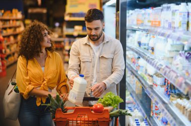 Süt seçen bir çift, modern bir marketin süt ürünleri bölümünde içi taze ürünlerle dolu bir alışveriş arabasını itiyor.