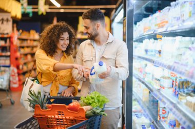 Genç bir çift markette süt seçiyor, sağlıklı bir yaşam için içi taze ürünlerle dolu bir alışveriş arabasını itiyorlar.