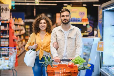 Genç, gülümseyen bir çift, marketten ellerinde taze yiyeceklerle alışveriş arabalarını itiyor. Sağlıklı yaşam tarzının ve birlikteliğin tadını çıkarıyorum.