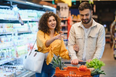 Market koridorunda yürüyen genç bir çift taze meyve ve sebzelerle dolu bir arabayı itiyorlar.