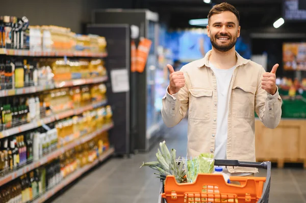 Mutlu sakallı adam bir süpermarket arabasını koridorda itiyor, geniş bir gülümseme içinde ve iki hevesli baş parmağını kaldırıyor.