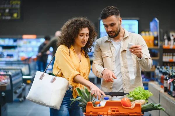 Genç bir çift, taze yiyecek seçerken fişe bakıyor ve modern bir markette alışveriş arabasını itiyor.