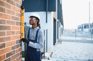 Kasketli ve güvenlik gözlüklü inşaat işçisi bir tuğla duvarın ruh seviyesiyle dikey hizasını kontrol ediyor.