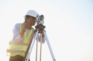 Siyah erkek mühendis çelik yelek ve şapka giyiyor. İnşaat alanında anket aleti ve telsiz kullanıyor.