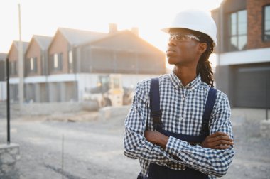 Kasketli, gözlüklü ve tulumlu genç siyahi bir adam inşaat sahasında güvenle duruyor.