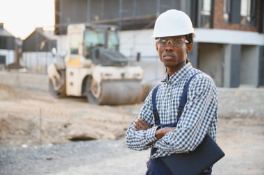 Afrika kökenli Amerikalı inşaat işçisi, kolları çapraz duruyor, inşaat sahasında güvenlik kıyafeti giyiyor.