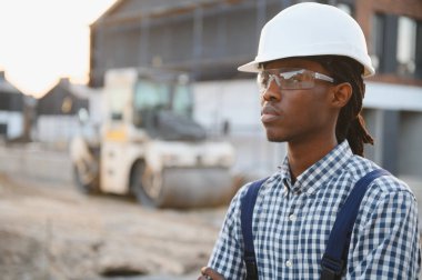 Siyah adam teknisyen, kasket ve güvenlik gözlüğü takıyor, yol inşaatında güvenle duruyor.