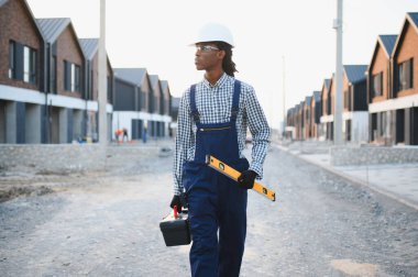 Erkek inşaat işçisi, sağlam bir şapka ve güvenlik gözlüğü takıyor, inşaat alanında alet çantası ve ruh seviyesi taşıyor.