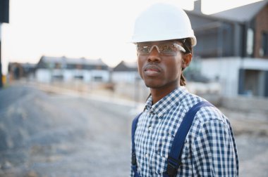 Sert şapka ve güvenlik gözlüğü takan Afro-Amerikalı genç bir adam konut inşaatında güvenle ayakta duruyor.