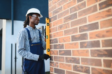 Siyah şapkalı ve güvenlik gözlüklü Afrikalı Amerikalı inşaat işçisi yeni inşa edilmiş tuğla duvarın dikey hizasını kontrol ediyor.