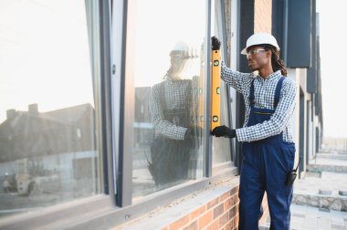 Afrikalı Amerikalı inşaat işçisi binanın ön cephesine yeni bir pencere yerleştiriyor, kesinlik için gerekli düzeyi sağlıyor.