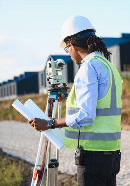 Siyah erkek arazi mühendisi planları kontrol ediyor, inşaat alanında kasket ve güvenlik yeleği giyiyor.