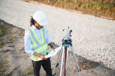 Sert şapka ve güvenlik yeleği giyen genç bir inşaat uzmanı yeni bir yol çalışması sahasının planlarını gözden geçiriyor.