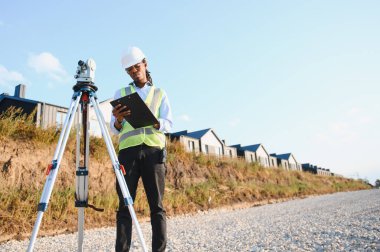 Jeodezi mühendisi sıkı bir şapka ve güvenlik yeleği giyiyor. Yeni konut alanındaki bir teodolit yakınındaki planları gözden geçiriyor.