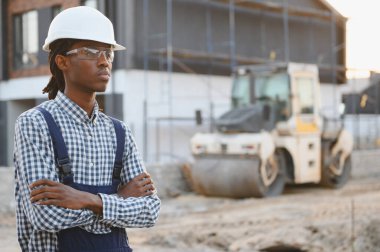 Güvenlik teçhizatlı genç siyah adam yeni bir inşaat projesinde kollarını kavuşturarak güven ve uzmanlık gösteriyor.