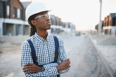 Kasket ve güvenlik gözlüğü takan genç siyah inşaat işçisi, inşaat alanında çapraz kollarla duruyor.