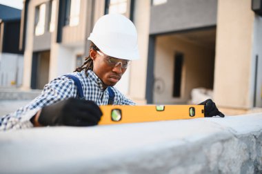 Genç siyah inşaat işçisi, bir inşaat alanının duvarındaki ruh seviyesini kullanarak, koruyucu gözlük ve şapka takıyor.