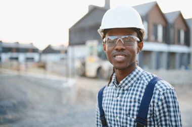 Genç Afrikalı Amerikalı adam, şapka ve güvenlik gözlüğü takıyor, modern bir inşaat alanında çalışırken gülümsüyor.