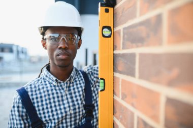 Genç siyahi inşaatçı güvenlik kıyafeti giyiyor ve ruhsal seviyesini tuğla duvarda kullanıyor. Kaliteli bir iş sağlıyor.