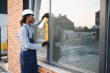 Afrika kökenli Amerikalı inşaatçı, pencere çerçevesini ruh seviyesine göre ayarlıyor, bir inşaat sahasında kurulum sırasında hassasiyet sağlıyor.
