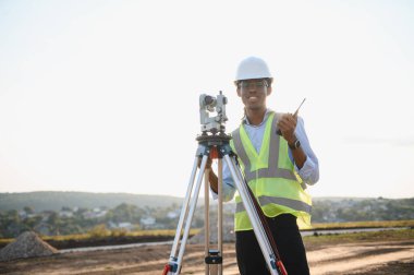 Gözetleme ekipmanları ve telsizle çalışan, güvenlik yeleği giyen ve inşaat alanında kasket takan bir arazi mühendisi.