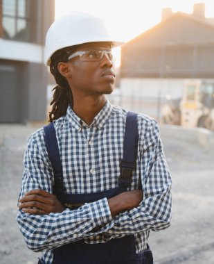 Siyah müteahhit koruyucu şapka ve güvenlik gözlüğü takıyor, kollarını kavuşturup inşaat alanına bakıyor.