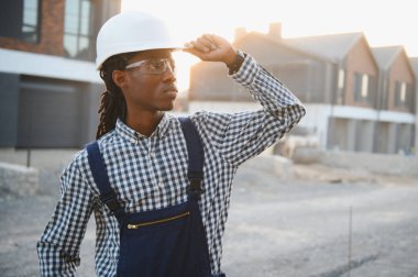 Afrika kökenli Amerikalı inşaat işçisi. Başlık ve güvenlik gözlüğü takıyor. İnşaat alanındaki inşaat çalışmalarını denetliyor.