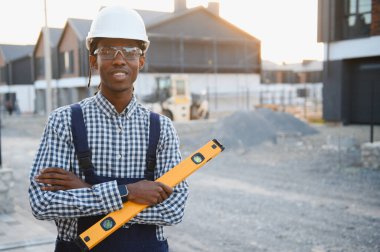 Sağlam şapka ve güvenlik gözlüğü takan kendine güvenen inşaat işçisi gelişmekte olan bir yerleşim bölgesinde ruh seviyesinde duruyor.