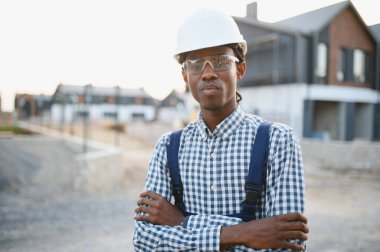 Afrika kökenli Amerikalı inşaat işçisi güvenlik ekipmanı takıyor, kolları çapraz duruyor, yeni bir yerleşim yerinin yanında.