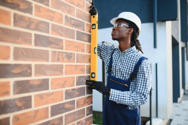 Kasketli ve güvenlik gözlüklü genç bir inşaatçı, ruhsal seviyeye sahip tuğla duvarı dikkatlice ölçüyor.