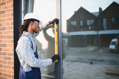 Başlık takan yetenekli genç bir inşaat işçisi yeni bir ev için yeni takılmış pencere çerçevesini kontrol ediyor.