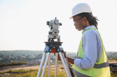 Sert şapka ve güvenlik yeleği giyen erkek arazi ölçümü ve mühendislik işi için teodolit kullanıyor.