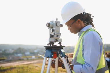 Siyah şapka ve güvenlik yeleği giyen Afrikalı Amerikalı erkek mühendis arazi taraması için tüm istasyonu kullanıyor.