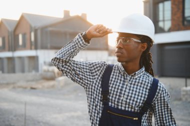 Afrika kökenli Amerikalı erkek inşaat işçisi, sıkı bir şapka ve güvenlik gözlüğü takıyor, gelişim sahasına bakıyor.