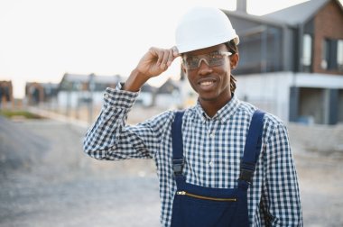 Gülümseyen Afro-Amerikan inşaat işçisi inşaat alanında dururken beyaz bir şapka ve güvenlik gözlüğü takıyor.