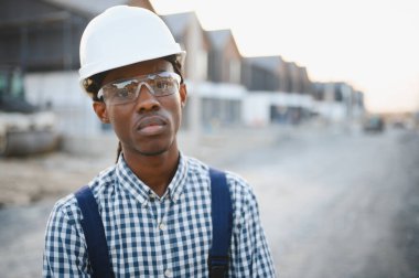 Siyah şapkalı ve güvenlik gözlüklü bir adam, bir inşaat alanında dikiliyor, ciddi ve odaklanmış görünüyor.