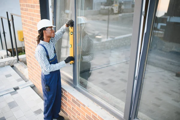 Sağlam şapka ve güvenlik gözlüğü takan yetenekli inşaat işçisi, yeni evdeki pencere kurulumu için ruh seviyesini kullanıyor.