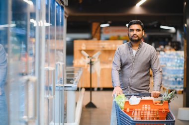 Market alışverişi yapan bir adam modern bir süpermarkette içi taze ürün ve malzemelerle dolu bir arabayı itiyor.