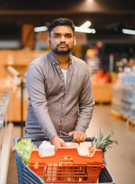 Market alışverişi yapan genç bir adam, modern bir perakende mağazası koridorunda içinde taze yiyecek olan bir arabayı itiyor.