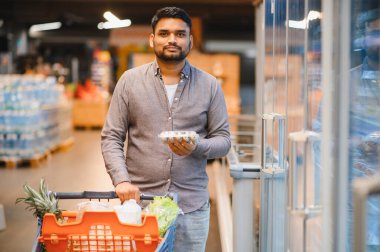 Genç adam alışveriş yapıyor, yumurta tutuyor ve markette alışveriş arabasıyla alışveriş yapıyor.