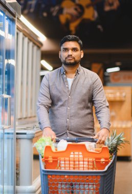 Kendine güveni tam bir alışveriş arabasını modern bir süpermarketten geçirip, taze ürünler ve tüketici malları alan bir adam.