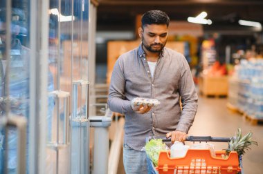 Genç adam modern bir süpermarkette alışveriş yapıyor, içi taze ürünlerle dolu bir arabayı itiyor, paketlenmiş bir şeyi dikkatlice kontrol ediyor.