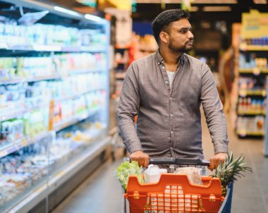 Taze ürün ve tüketici ürünleriyle dolu alışveriş arabalarını iten adam, bir market koridorunda geziniyor.