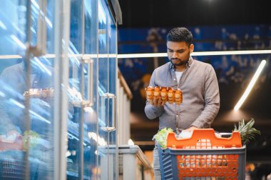 Bir karton yumurtayı elinde tutan bir adam, markette onlara bakıyor, bir alışveriş arabası dolusu ürünle.